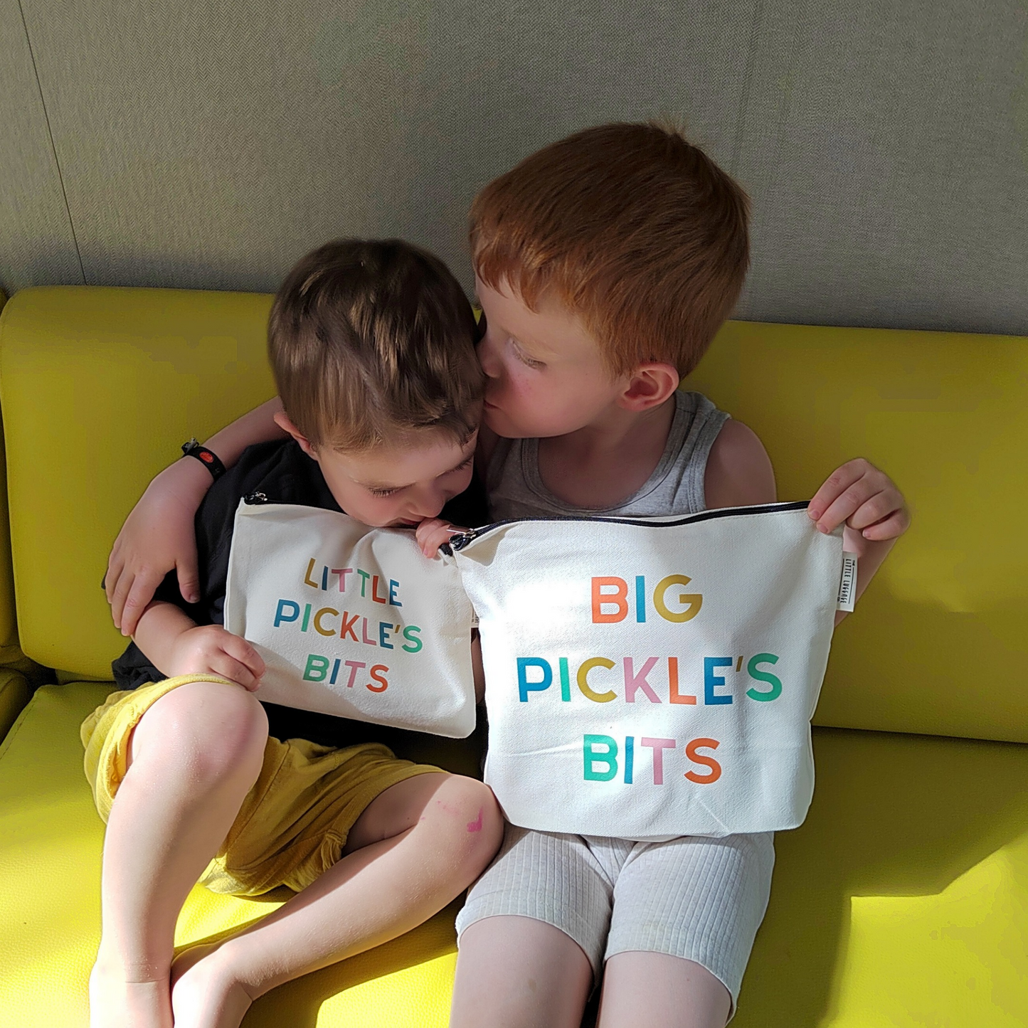 Two children sitting on a yellow couch with 'Little Pickles Bits' and 'Big Pickles Bits' bags.
